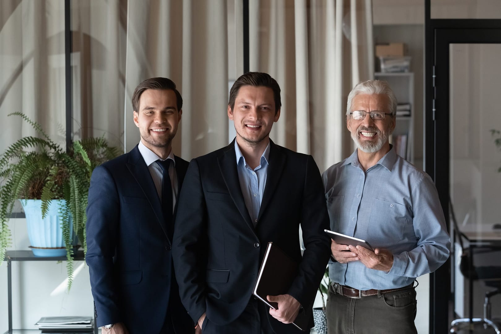 Three smiling men of different ages in business attire, standing together in an office.